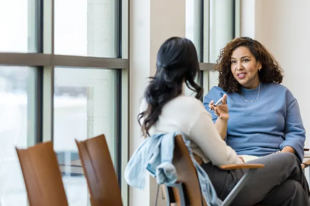 Two adults talk sincerely while sat in private on two chairs in a canteen setting
