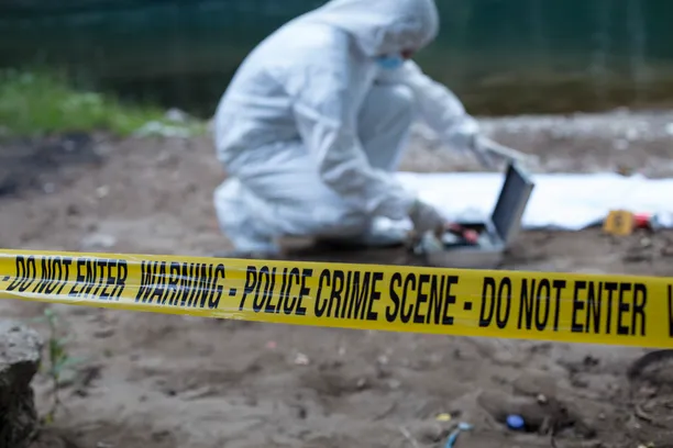 A crime scene investigator dressed in full white overalls looks for tools in a silver case at an active crime scene near a river cordoned off by yellow crime scene tape