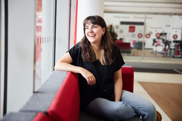 Postgraduate Research English student Joanne Spearman laughs as they sit and lean on the back of a grey and red sofa while looking out the window in the foya of the South Wales Business School
