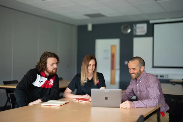 A group of three sport researchers, including PhD students and doctoral supervisors, sit around a table chatting with notepads and a laptop