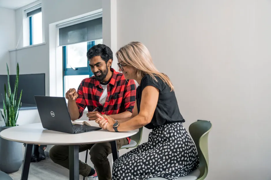 Two colleagues are working together, making notes and looking at a laptop.