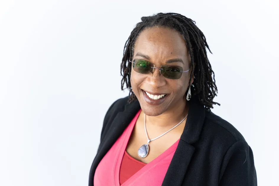 Sociology professor Dr Roiyah Saltus smiles at the camera while wearing sunglasses in front of a white backdrop