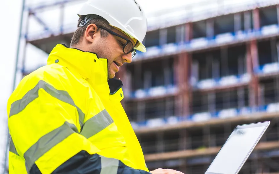 A worker with a yellow high visibility jacket and a white hard hat looks at their laptop at a construction site