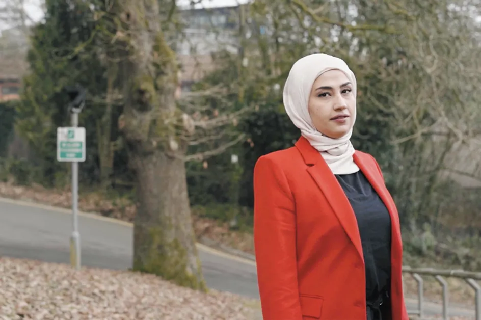 Syrian graduate Zaina Aljumma looks at the camera side-on while wearing a read coat in front of autumnal looking trees in a park