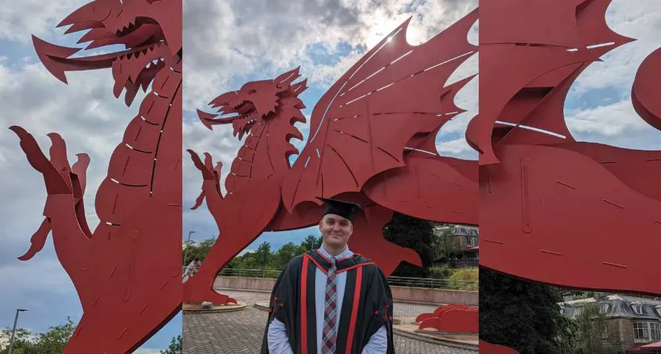 Graduate James McFarlane dressed in a suit, graduation robes and a graduation hat proudly stood in front of the big red dragon outside Newport's ICC