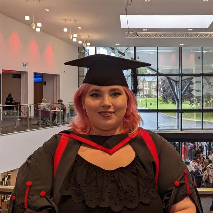 Mae Jess James, un o raddedigion, yn gwisgo gwisg graddio a chap graddio wrth wenu ar y camera o'r balconi yn ICC Casnewydd gyda ffenestr y cyntedd y tu ôl iddynt