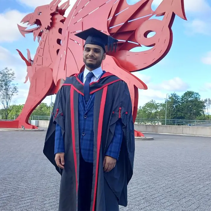 Graddedig Shafiul Mannan yn gwisgo gwisg graddio a chap graddio o flaen y ddraig goch y tu allan i ICC Casnewydd