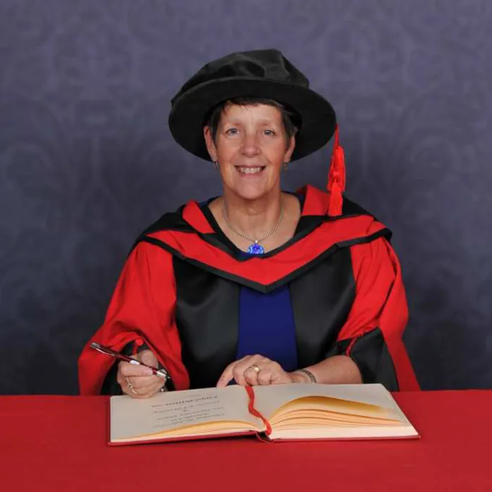 Pippa Britton anrhydeddus graddio yn eistedd wrth fwrdd coch mewn gwisgoedd graddio a het raddio gyda beiro a llyfr