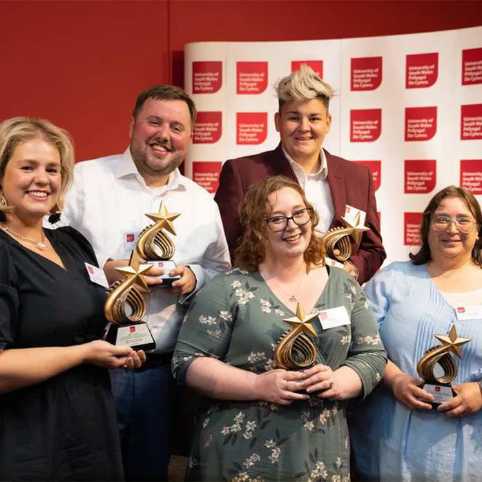 Olivia Sills, Timothy Raggett, Bethan Howell, Deborah Crockett and Lucinda Coultas holding star-shaped trophies in front of a white backdrop with USW logo tiles and a red wall