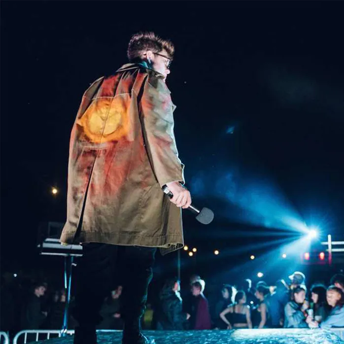 An actor facing away from the camera on an outdoor stage dramatically lit in the background