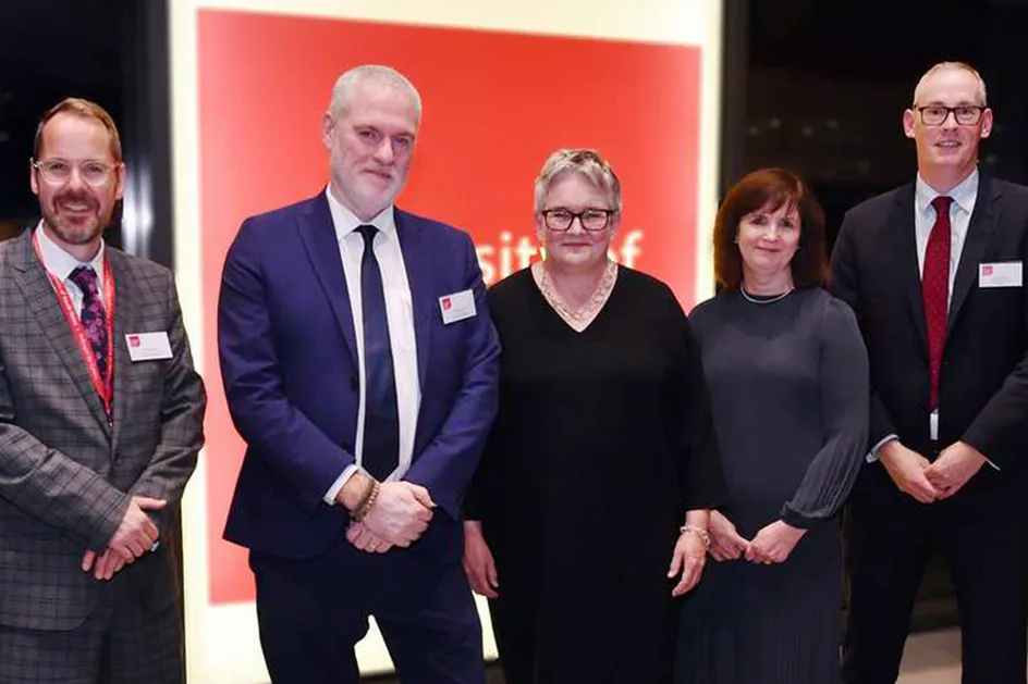 Jared Davies, Ben Calvert, Helen Brand OBE, Nicola Gilbert, a Lloyd Powell yn gwenu ar y camera yn sefyll o flaen logo Prifysgol De Cymru