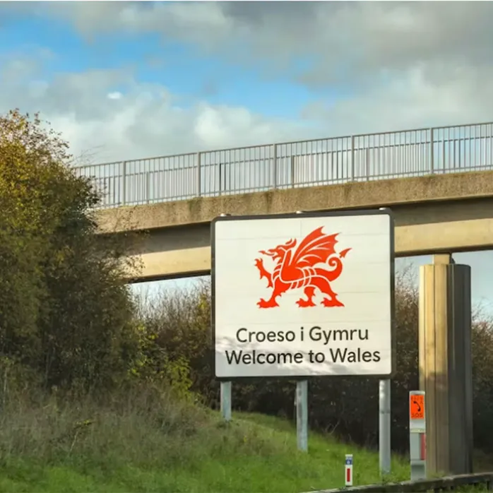 Arwydd ffordd Croeso i Gymru wedi’i  amgylchynu gan goed ar ochr ffordd