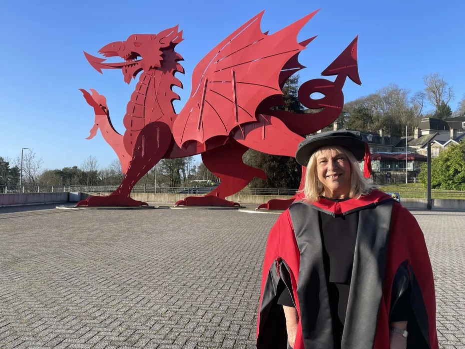 Menyw yn gwenu yn gwisgo gŵn graddio du a choch a het. Mae hi'n sefyll o flaen draig fetel goch fawr ac mae'r awyr yn las llachar.
