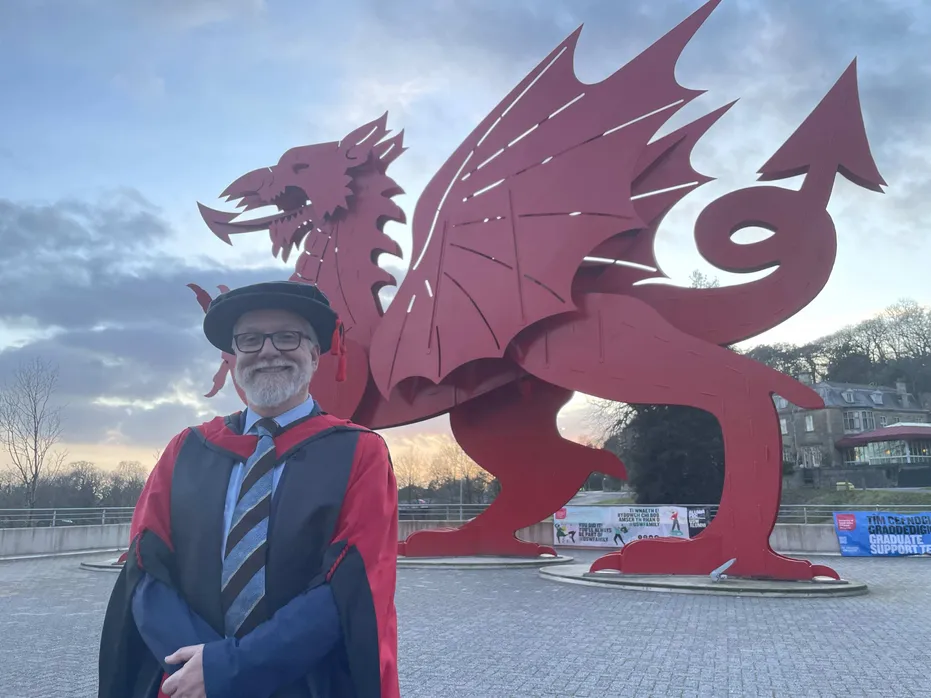 Dyn yn gwenu yn gwisgo gŵn graddio du a choch a het. Mae hi'n sefyll o flaen draig fetel goch fawr.