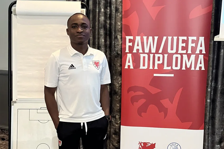 Mae Eboboritse yn gwisgo crys-t gwyn ADIDAS gyda logo draig goch Cymdeithas Bêl-droed Cymru arno. Mae e'n sefyll wrth ymyl baner sy'n dweud 'FAW/UEFA A DIPLOMA'.