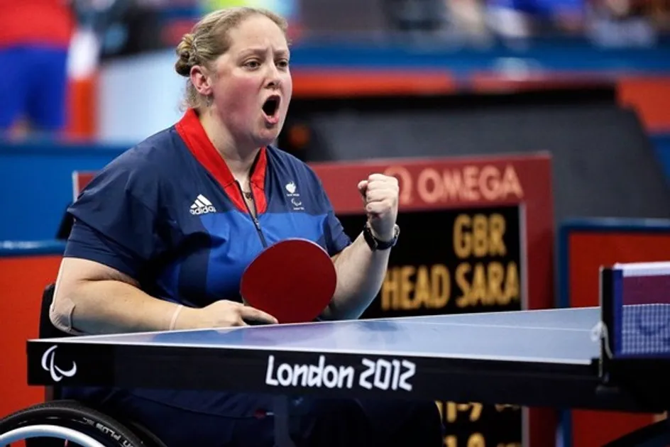 Mae Sara yn eistedd mewn cwrt tennis bord yn ei chadair olwyn. Mae hi'n dal bat tennis bord mewn un llaw ac yn dyrnu'r awyr i ddathlu gyda'i llaw arall. Mae gan y bwrdd logo 'Llundain 2012' arno.