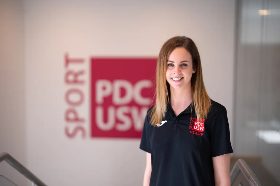 Mae Hannah yn gwisgo crys-t du gyda logo coch Chwaraeon PDC arno. Mae ganddi wallt hir tywyll, syth ac mae'n gwenu ar y camera. Y tu ôl iddi mae logo mawr coch Chwaraeon PDC ar y wal.