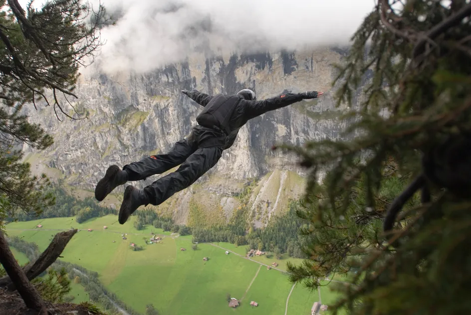 A person in a black wingsuit is jumping off a cliff, arms outstretched, with a scenic mountain landscape in the background. The valley below features green fields, small houses, and a winding road, while the cliff face is partially shrouded by clouds. Pine trees frame the sides of the image.