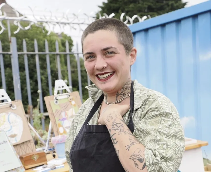 A smiling person with a buzz-cut hairstyle is standing outdoors in front of an art display. They are wearing a light green patterned shirt and a black apron. The person has visible tattoos on their neck and arms, including a prominent tattoo of a straight razor on their forearm. They are also wearing a nose ring and a necklace.
