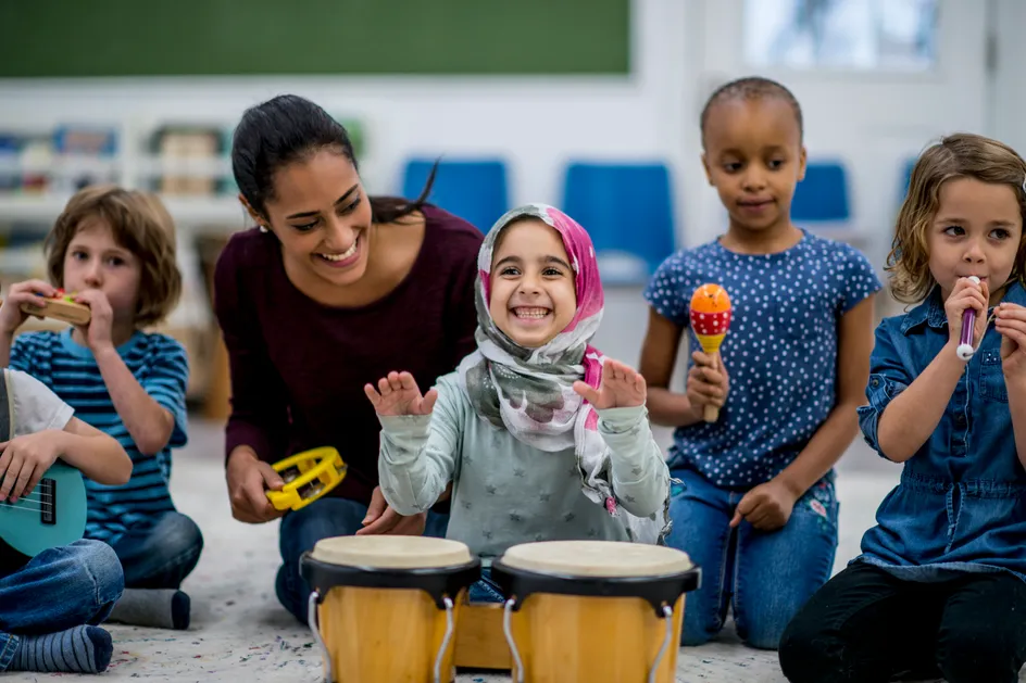 A group of young children and an adult are joyfully playing musical instruments together in a classroom. The children are using instruments such as drums, maracas, a small flute, and a ukulele. The adult, sitting in the centre, is smiling and holding a tambourine, engaging with the children. One child wearing a colourful hijab is happily playing the drums, smiling brightly. The setting is bright a