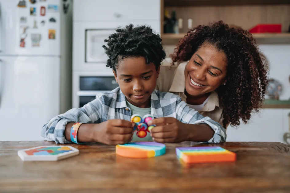 A young child sits at a kitchen table, focused on playing with colorful sensory toys, including a fidget spinner and pop-it. An adult, likely a parent or caregiver, leans in smiling supportively, sharing a moment of encouragement and connection.