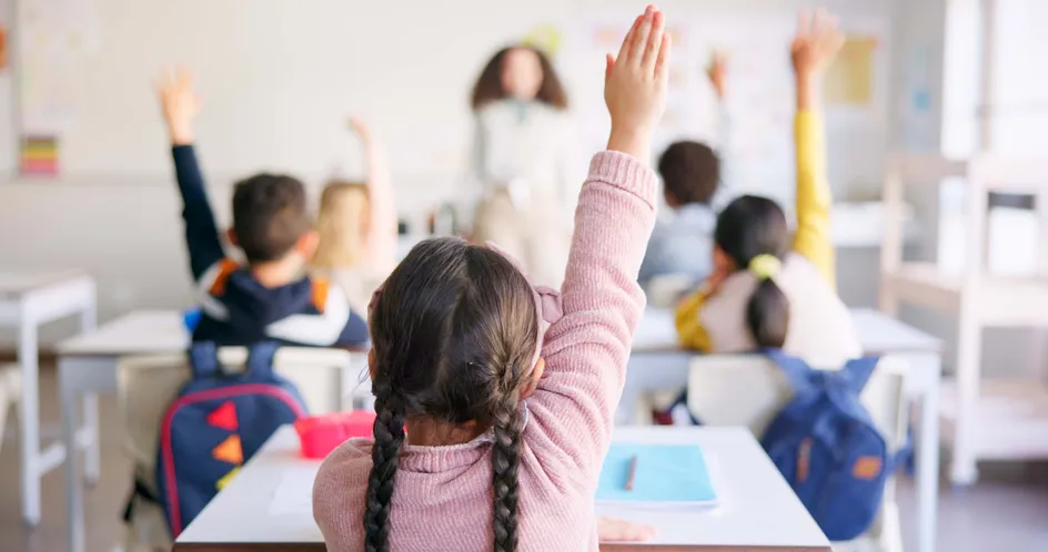 A classroom with young students sitting at desks, facing the teacher at the front. Several children have their hands raised.