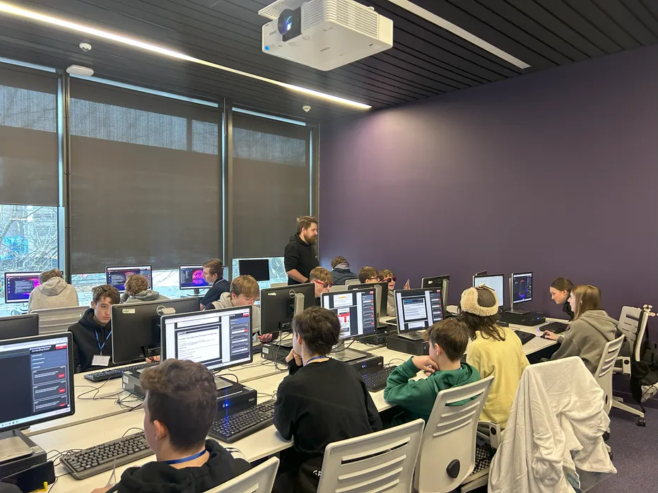 Students sit at computers, engrossed in a cyber security activity. An industry partner stands up in the background of the image, assisting two students with the activity.