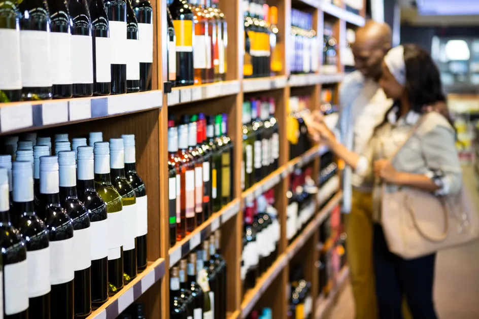 A couple browsing the wine aisle in a shop, with shelves of various wine bottles in the foreground.