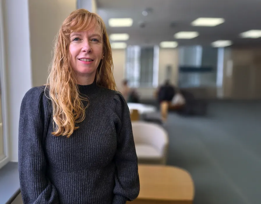 Katy has long, wavy red-blonde hair wearing a dark grey jumper. She is smiling and looking at the camera. The background shows a bright office setting with blurred furniture and windows.
