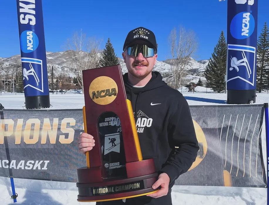 Sam Thomas, wearing sunglasses and a University of Colorado Ski Team cap, holds the NCAA National Champion trophy while standing in front of a snowy backdrop with banners reading 'NCAA Skiing' and 'Champions.'