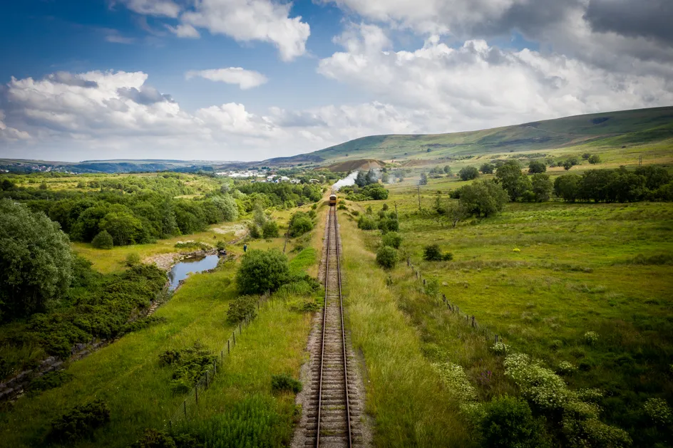 A scenic view of a railway track cutting through a lush green countryside with rolling hills in the background. A train is visible in the distance.