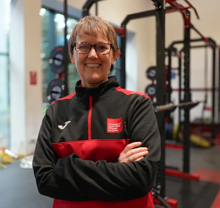 Denise has short, light brown hair and glasses. She stands with her arms crossed in a gym. She is wearing a black and red University of South Wales sports jacket with the University logo on the chest. Behind her are weightlifting racks, barbells, and other gym equipment.