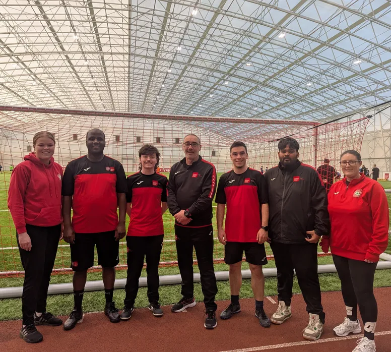 A group of seven people is posing in front of a football goal inside a large indoor sports facility with a high, transparent ceiling. They are wearing matching red and black sportswear, with some in jackets and others in short-sleeved shirts. The group consists of men and women of diverse backgrounds, all smiling at the camera. The artificial turf field behind them has other people engaging in foo