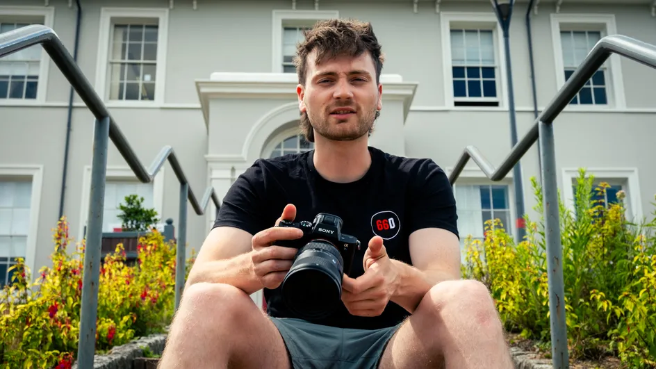 Mae dyn ifanc yn eistedd ar risiau awyr agored o flaen adeilad gwyn mawr, yn dal camera Sony. Mae'n gwisgo crys-t du gyda logo 