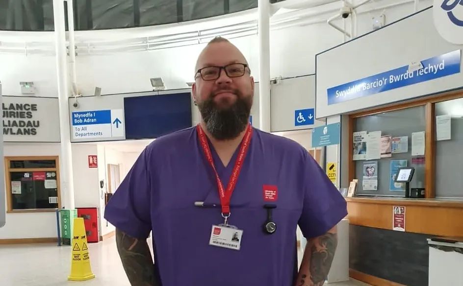 Owen Nash, an adult nursing student, stands in a hospital reception area wearing purple scrubs and a red lanyard with a University of South Wales ID badge. He has tattoos on both arms, glasses, and a beard, smiling with his hands in his pockets.
