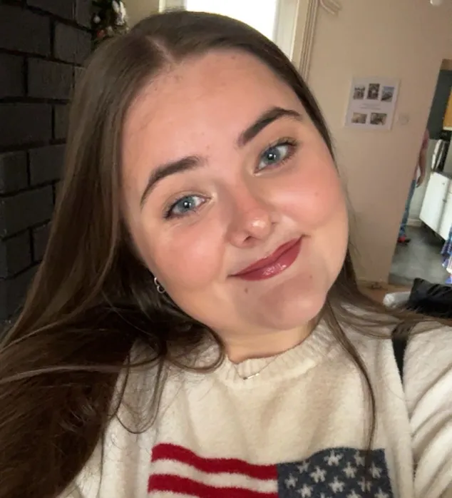 A young woman with long brown hair is smiling at the camera in a cozy indoor setting. She is wearing a cream-colored sweater featuring a large American flag design on the front.