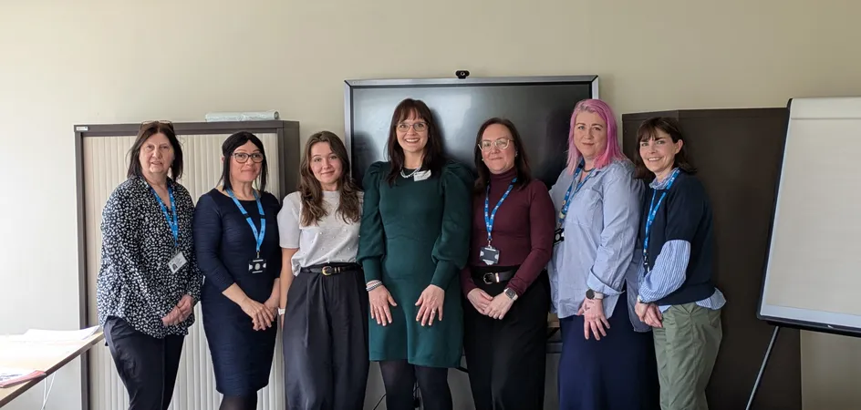 A group of seven women standing side by side in a meeting room, all wearing ID badges on blue lanyards. They are smiling at the camera in front of a large screen and filing cabinets.
