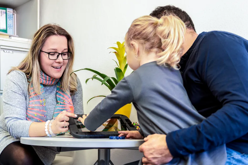 A health visitor smiles while interacting with a young girl and her father. The child is using a tablet device on the table, with both the health visitor and child engaged in the screen.