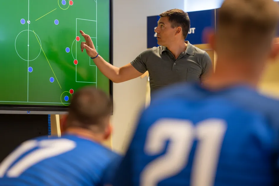 A coach in a grey polo shirt points at a digital tactics board displaying a soccer field with player positions and movement lines, while two players in blue jerseys with numbers on their backs listen attentively.