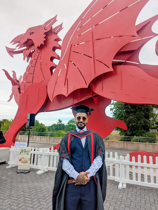 A man wearing a graduation cap and gown stands in front of a large red dragon sculpture. He is dressed in a dark suit with a red-accented academic robe and tie, wearing sunglasses, and has tattoos on his hands.