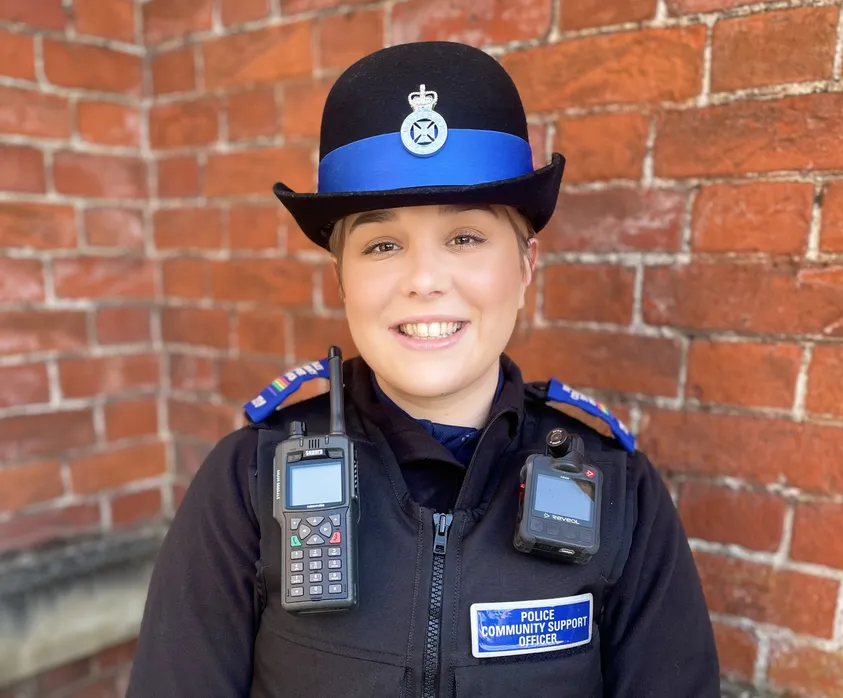 A smiling Police Community Support Officer stands in front of a red brick wall. She is wearing a black uniform with a name badge reading 