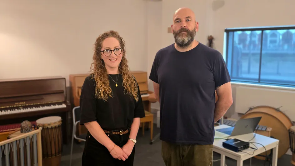 A woman with long curly hair, glasses, and a black outfit stands smiling next to a man with a shaved head, a full beard, and a dark t-shirt. They are in a music room with instruments visible, including a piano, drums, and a laptop on a table in the background.