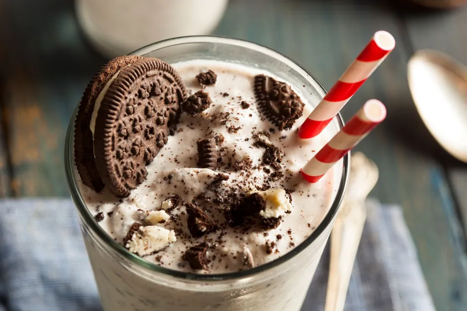 A cookies and cream milkshake sits on a wooden table in a tall glass with a spoon beside it. There is a large biscuit in the milkshake along with crumbled chocolate cookies. There are two red-and-white straws standing in the glass.