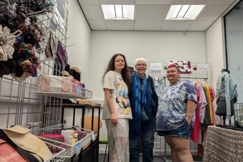 Three talented makers currently selling in the shop, smiling together in a small space filled with fabric items, bags, and upcycled clothing displayed on wire racks.