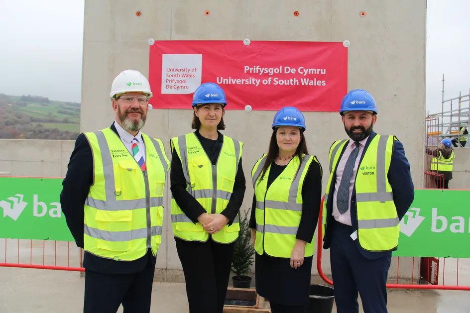 Four individuals wearing high-visibility vests and hard hats standing in front of a concrete wall with a red University of South Wales banner during the Calon topping out ceremony.