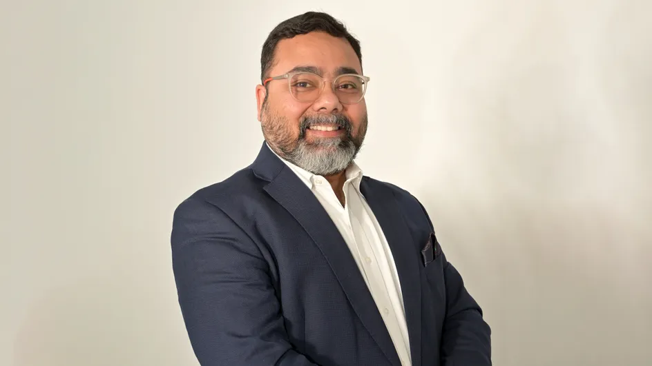 A person wearing a dark blazer over a white shirt, standing against a plain light-coloured background.