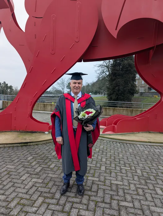 A smiling man wearing a graduation cap and a grey academic gown and holding a bouquet of flowers.