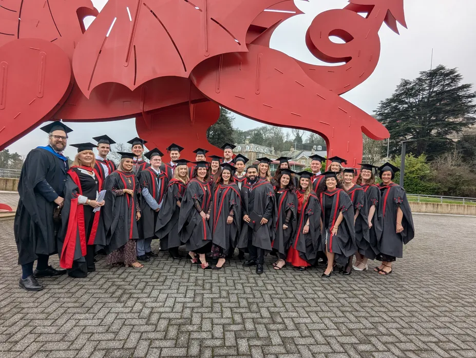 A large group of graduates wearing black gowns and mortarboards stand smiling for a group photo beneath a large red dragon sculpture.