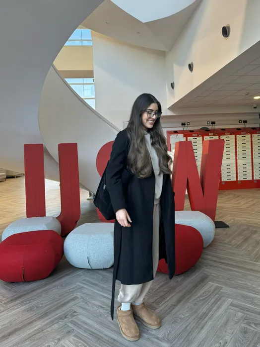 A woman with long dark hair and glasses stands indoors in a modern atrium with curved white architecture. She wears a long black coat over a beige outfit and tan ankle boots, smiling while looking slightly downward.