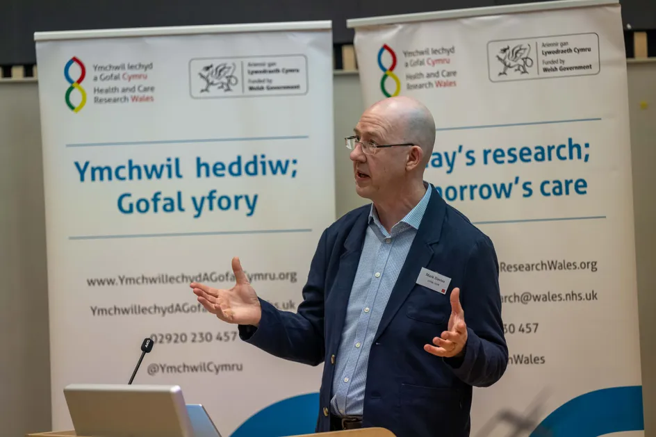 Mae dyn mewn blazer glas yn siarad wrth lecsenn, gan wneud symudiadau gyda'r ddwylo, o flaen baneri Ymchwil Iechyd a Gofal Cymru sy'n dangos testun Cymraeg ac Saesneg.